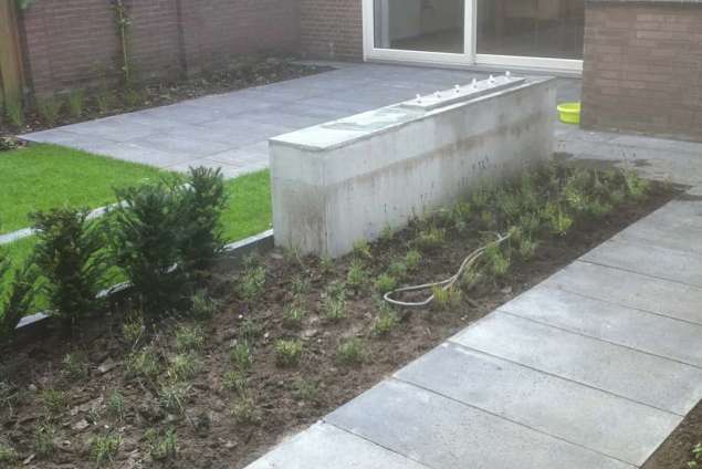 strak waterelement in een betonmuur een bak van rvs met daarin 6 waterspuwers. in de tuin liggen schellevis tegels 100x50x5 antrasiet  tuin aanleggen in de wijk de blaak tilburg hovenier